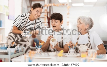 Enthusiastic senior female teacher sharing experience pottery craftsmanship, assisting happy young guy and girl, painting ceramic mugs and plates at table in workshop Royalty-Free Stock Photo #2388467499