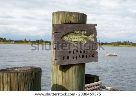 A wooden sign posted on a pier or wharf warning people to please no fishing in the area. The wooden signage has a graphic of a fish and black text. There's water and a boat in the background.