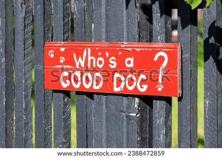 A vibrant red wooden sign with a funny saying: who's a good dog? with white painted letters. The fence barricades a lush green grassy garden. The black wooden fence is made of narrow sticks.  