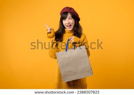 Young Asian woman, wearing a yellow sweater and red beret, indulges in a shopping extravaganza and pointing finger to free copy space against a vibrant yellow backdrop.