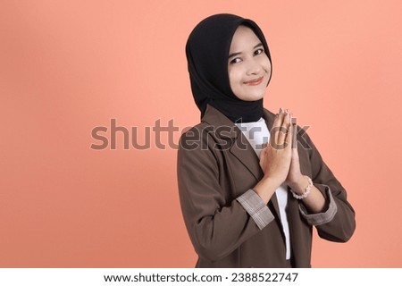 Smiling young business woman 
 Asian Muslim woman gesturing Eid Mubarak greeting company wearing brown suit isolated over peach background