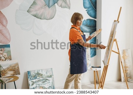 Sideways profile elderly fun satisfied artist woman 50 years old wear casual clothes standing near easel with painting artwork paint spend free spare time in living room indoor. Leisure hobby concept