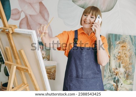 Elderly artist woman 50 years old wears casual clothes stand near easel with painting artwork paint talk speak on mobile cell phone spend free spare time in living room indoor. Leisure hobby concept