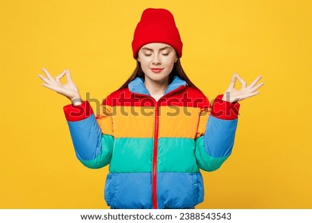 Young spiritual woman wear padded windbreaker jacket red hat casual clothes hold spread hands in yoga om aum gesture relax meditate try calm down isolated on plain yellow background. Lifestyle concept
