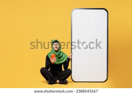 Full body young asian woman wear green hijab abaya black clothes point on big huge blank screen mobile cell phone use smartphone isolated on plain yellow background. People uae islam religious concept