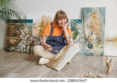 Full body happy smiling fun cheerful elderly artist woman 50 years old wears casual clothes sit on floor painting on canvas artwork spend free spare time in living room indoor. Leisure hobby concept