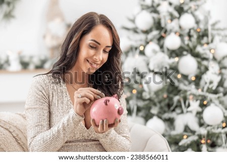 Smiling woman puts a coin into a pink piggybank sitting next to a Christmas tree.