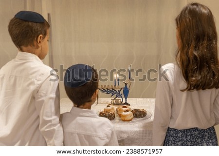 Three Jewish children looking at the Menorah with 8 lit burning candles for Jewish Hanukkah holiday on table at home. Celebrating Chanukah festival of lights. Dreidel and Sufganiyot donuts on a plate.