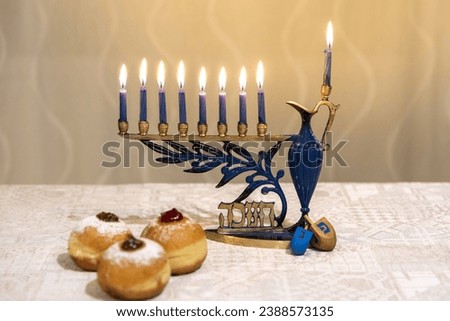 Menorah (Chanukkia) with 8 lit burning candles for Jewish Hanukkah holiday on table at home. Celebrating Chanukah festival of lights. Dreidel and Sufganiyot donuts sweet cultural food on a plate.