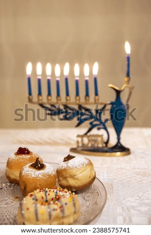 Sufganiyot donuts sweet cultural food on a plate, in focus . Menorah (Chanukkiah) with 8 lit burning candles for Jewish Hanukkah holiday on table at home, in the background