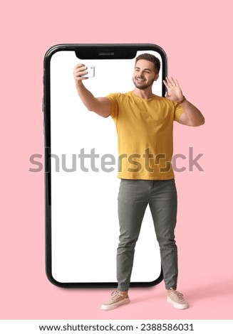 Young man taking selfie and big smartphone on pink background