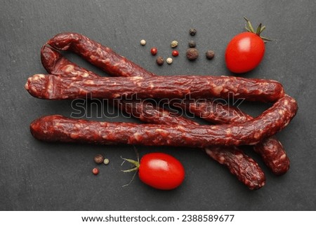 Many thin dry smoked sausages, peppercorns and tomatoes on black table, flat lay Royalty-Free Stock Photo #2388589677