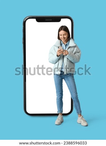 Pretty young woman in warm clothes and big smartphone on light blue background
