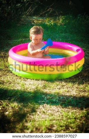A cute little baby boy is playing with toys while bathing in a little colorful wading pool on meadow garden field outdoor. Summer time. Joy and happiness. Love and family  Royalty-Free Stock Photo #2388597027
