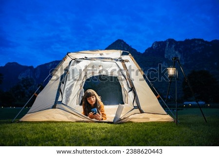 Charming asian woman with beautiful smile reading good news on mobile phone while lying in tent and warm night light under dark blue sky twilight time. The tent lights at night from the lamp camping. 