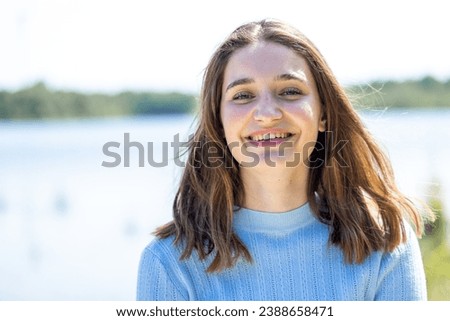Amidst a natural backdrop, a pretty girl with open eyes and a beaming smile exudes cheerfulness. Her long brown hair and trendy accessories complement the organic beauty surrounding her, creating a Royalty-Free Stock Photo #2388658471