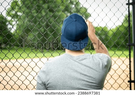 A man in a grey t-shirt and blue baseball cap is seen from behind, leaning on a chain-link fence, looking onto a sports field