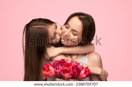 Little girl kissing cheerful woman in cheek presenting bouquet of tulips for spring holiday on pink background