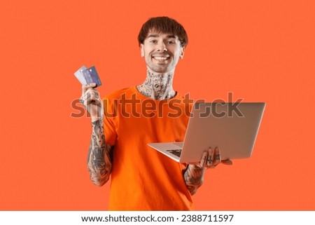 Tattooed young man with credit cards and laptop on orange background
