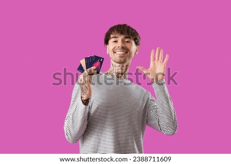 Tattooed young man with credit cards waving hand on purple background