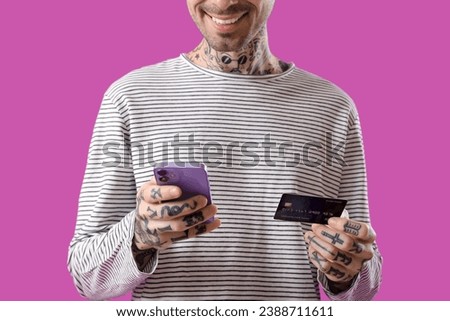 Tattooed young man with credit card using mobile phone on purple background, closeup