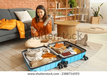 Young woman with mobile phone and thermos packing winter clothes in suitcase at home Royalty-Free Stock Photo #2388716593