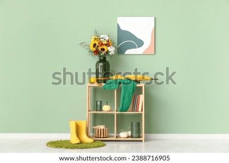 Shelving unit with bouquet of autumn flowers, pumpkins and umbrella near green wall