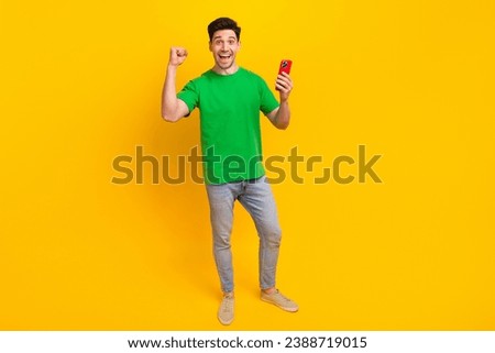 Full size photo of impressed guy wear stylish t-shirt denim pants hold smartphone win gambling isolated on yellow color background