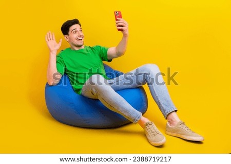 Full size photo of positive guy wear t-shirt sit on bean bag waving palm at smartphone on video call isolated on yellow color background