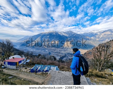 Male trekker hiker standing looking down at village houses and himalaya mountains in distance and cloudy sky in manali, kullu, shimla, kedarnath in India Royalty-Free Stock Photo #2388736961