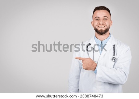 Cheerful male doctor in white coat pointing to the left at free space with stethoscope around his neck, suggesting direction or choice against light grey background