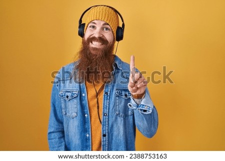 Caucasian man with long beard listening to music using headphones showing and pointing up with finger number one while smiling confident and happy. 