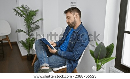 Relaxed young hispanic male settling into a waiting room chair, engrossed in a book - a handsome portrait of rest, focus, and ongoing pursuit of knowledge. indoor corridor forms a serene background. Royalty-Free Stock Photo #2388755523