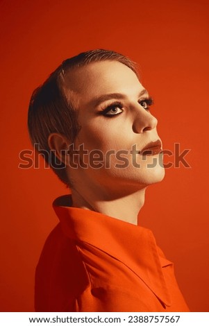 Extravagant young man with stylish hairstyle and wright makeup, lipstick and lashes posing against red studio background. Concept of male makeup, fashion, lgbtq community, self-identity, acceptance