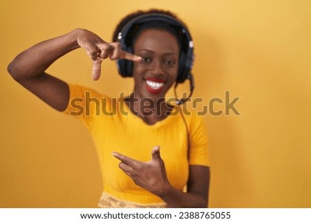 African woman with curly hair standing over yellow background wearing headphones smiling making frame with hands and fingers with happy face. creativity and photography concept. 