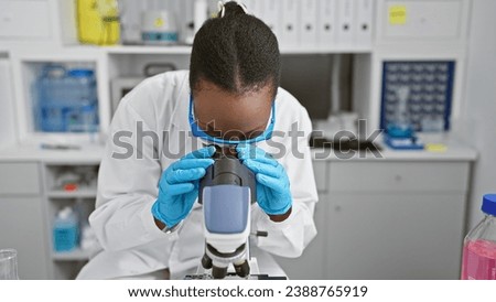 Focused african american woman scientist engrossed in scientific research, on microscope analysis in a bustling lab; her glasses symbolizing a quest for medical discovery.
