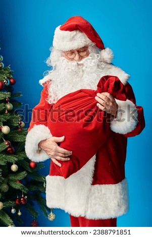 Santa Claus with beard and eyeglasses looking at red sack bag with Christmas presents on blue