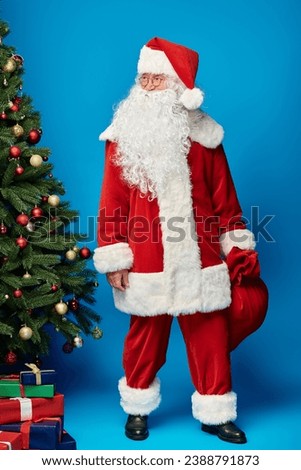 happy Santa Claus with beard and eyeglasses holding sack bag with presents near Christmas tree
