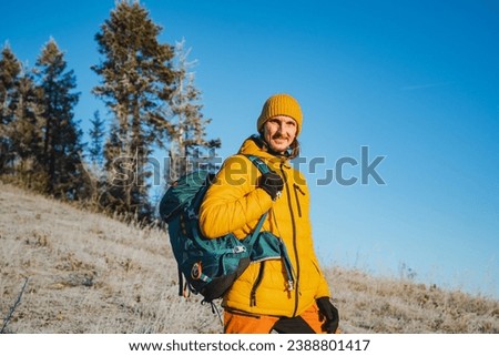 Tourist guy with backpack standing against blue sky background, male traveler smiling at camera, man in yellow jacket. Trekking in the mountains. High quality photo
