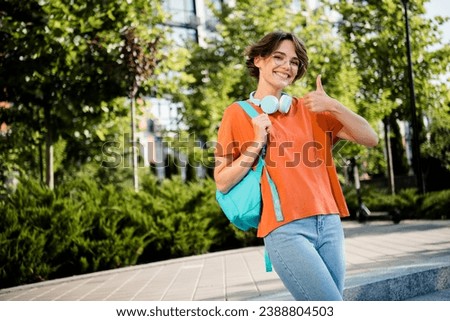 Photo of good mood cheerful lady wear orange t-shirt spectacles rucksack showing thumb up outside urban city street