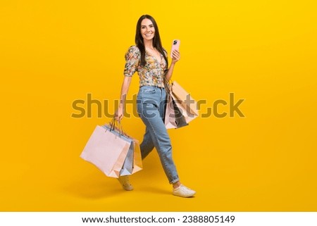 Full size photo of lovely woman wear top denim pants go empty space hold shopping bags smartphone isolated on yellow color background