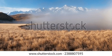 Beautiful sunny foggy morning in the mountains. Autumn frost on dry grass. Snowy peaks. North Chuysky Range and Lake Dzhangyskol. Russia, Altai Republic. Panoramic view. 