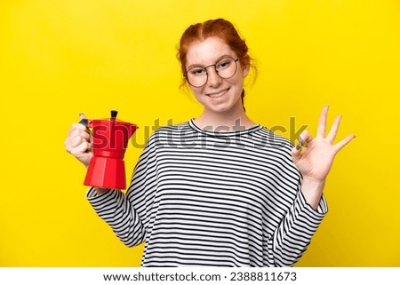 Young reddish woman holding coffee pot isolated on yellow background showing ok sign with fingers