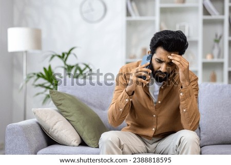 Angry angry man talking on the phone at home, dissatisfied customer sitting on the sofa in the living room yelling at the interlocutor. Royalty-Free Stock Photo #2388818595