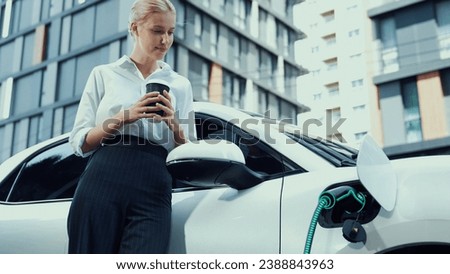 Businesswoman drinking coffee after recharge her electric car from charging station at residential parking car. Futuristic eco-friendly EV car use alternative energy in city urban lifestyle. Peruse