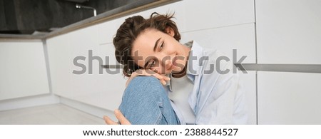 Portrait of brunette girl daydreaming, sitting on kitchen floor at home and smiling, looking aside with dreamy, thoughtful face expression.