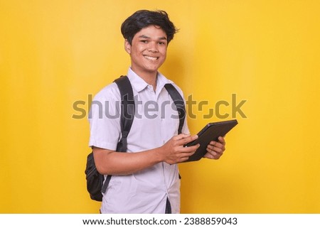 Portrait of smart young Asian male student in school uniform looking at camera smiling while holding tablet smartphone over yellow background