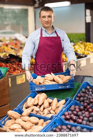 Portrait of successful greengrocery shop owner displaying fresh organic produce on counter, holding box of sweet potatoes and smiling confidently at camera....
