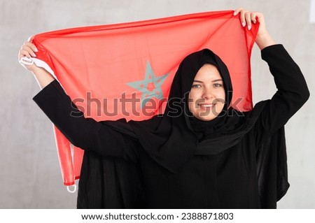 Smiling joyful Muslim woman in traditional black hijab holding Moroccan flag. Portrait of Muslim woman holding Moroccan flag on gray background