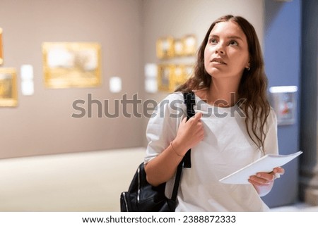 Portrait of a young girl visitor with a paper guide at the exhibition of paintings in the museum.. Royalty-Free Stock Photo #2388872333